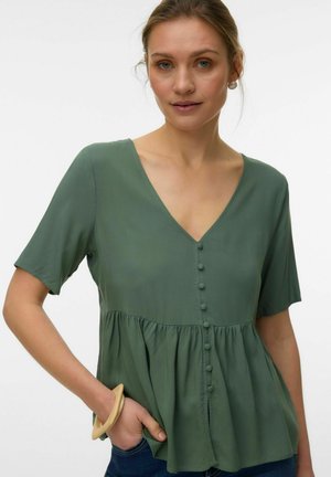 Kobieta ubrana w zieloną bluzkę z dekoltem w serek, z materiałowymi guzikami, krótkimi rękawami i marszczoną talią, z ręką w kieszeni dżinsów i drewnianą bransoletką.