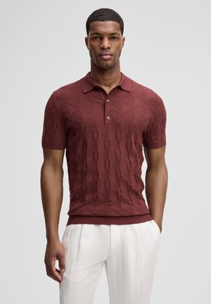 Mann trägt ein strukturiertes, kurzärmliges Poloshirt in Burgunderrot und weiße Hose, steht vor einem schlichten hellen Hintergrund.