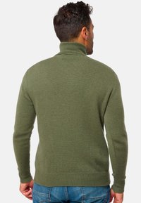 Sweatshirt col roulé vert en maille côtelée avec une coupe ajustée et des manches longues, fabriqué à partir d'un matériau tricoté texturé. Porté sur un jean bleu.