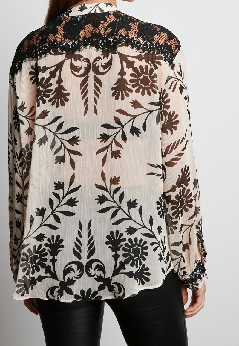 Blouse longue transparente à manches longues avec motifs floraux noirs et détail en dentelle noire sur le haut du dos et les poignets, portée avec un pantalon noir.