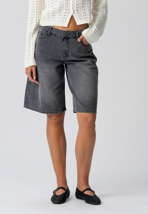 Person iført sorte denim Bermuda-shorts, en hvid strikket cropped sweater og sorte Mary Jane-sko, stående mod en lys baggrund.
