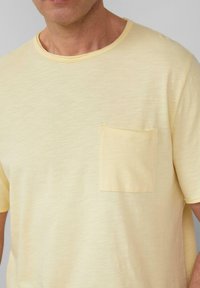 Gele katoenen t-shirt met ronde hals en korte mouwen, voorzien van een klein zakje op de linkerborst. De textuur lijkt zacht en licht van gewicht.
