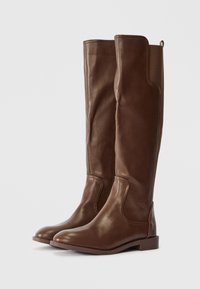 Botas de couro castanho até ao joelho com textura suave, bicos arredondados e um salto baixo em bloco. Apresentam costura em contraste e painéis laterais.