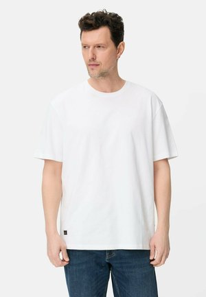 Mand med kort krøllet hår iført en ensfarvet hvid kortærmet t-shirt og blå jeans, der ser til sin venstre side mod en hvid baggrund.