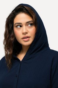 Dunkelblaue Kapuzenbluse aus strukturierter Fabric, mit einem durchgeknöpften Vorderteil und einem entspannten Schnitt. Akzente sind die sichtbare Kapuze und der Kragen.