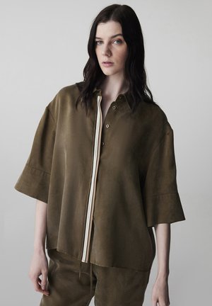 Jonge vrouw met donker haar draagt een loszittend olijfgroen overhemd met korte mouwen en bijpassende broek tegen een effen lichte achtergrond.