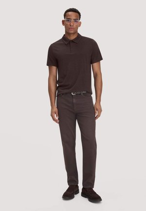 Mann mit transparenten Brillen, dunkelbraunem kurzärmeligem Poloshirt, passenden Hosen und dunklen Schuhen, der vor einem einfarbigen Hintergrund steht.