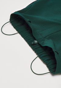 Dark green fabric bag with elasticated top, drawstring closure, and toggle. Smooth texture with a gathered design. Simple and functional.