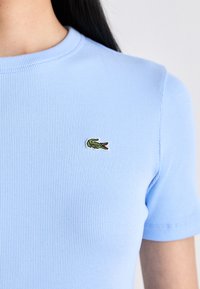 T-shirt à manches courtes bleu clair à côtes avec un col rond. Présente un petit logo de crocodile vert brodé sur le côté gauche de la poitrine.
