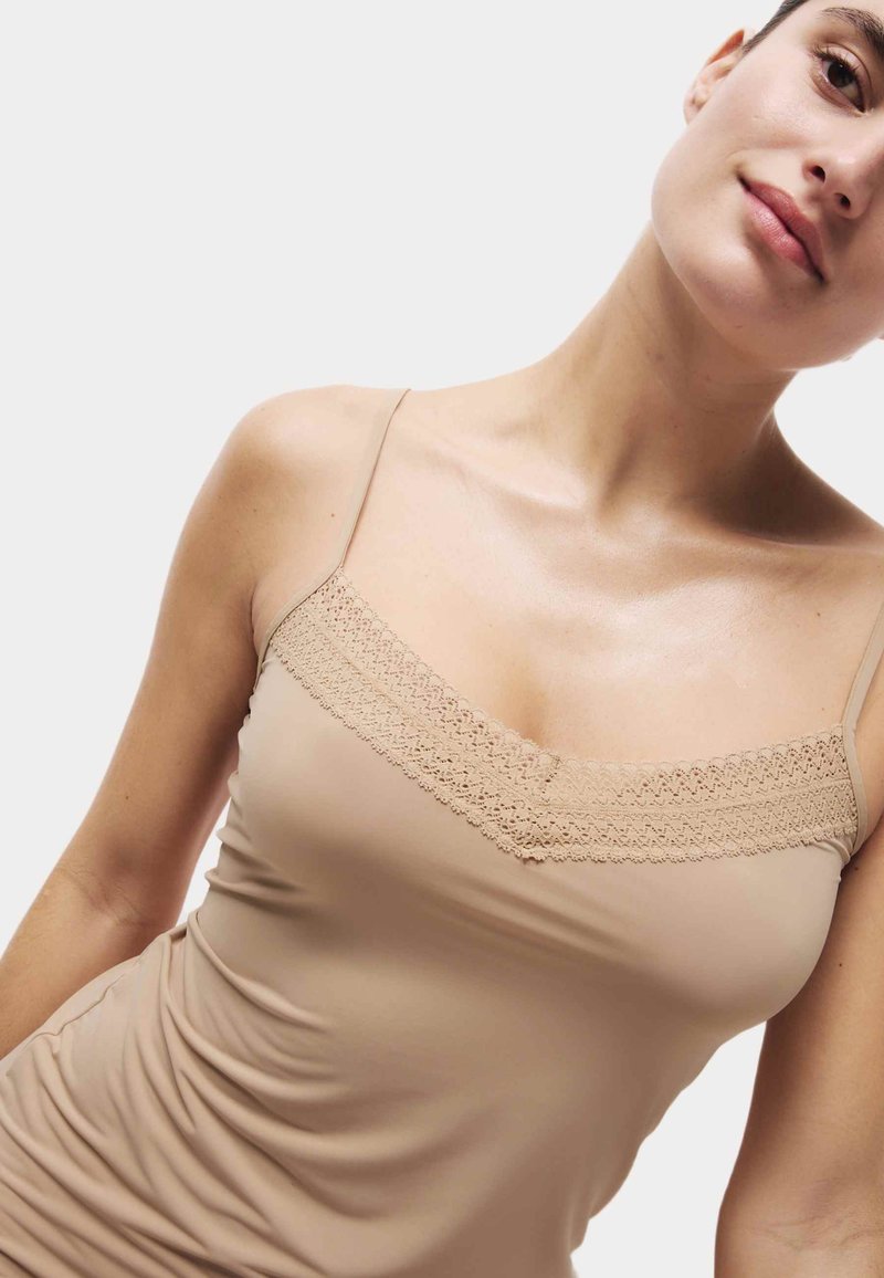 Nude tanktop met dunne bandjes en kanten rand bij de halslijn. Glad, rekbaar materiaal met een aangesloten silhouet.