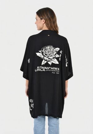 Mujer con una chaqueta estilo kimono negra con diseños de rosas blancas y texto japonés, combinada con vaqueros azul claro, de pie mirando hacia atrás.