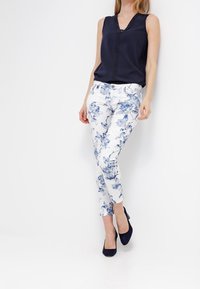 Blusa sin mangas azul marino con escote en V y pantalones estampados de flores en blanco y azul. Modelo llevando tacones azul marino, de pie frente a un fondo blanco.