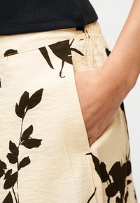 Main dans la poche d'une jupe beige à imprimé floral noir, associée à un haut côtelé noir.