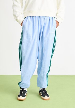 Persoon draagt lichtblauwe Adidas trainingsbroek met groene strepen, zwart-witte sneakers en een crème sweater met handen in de zakken.