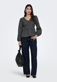 Femme debout portant un chemisier à carreaux foncé à manches longues, un jean large foncé, des talons noirs, tenant un sac noir dans sa main gauche.