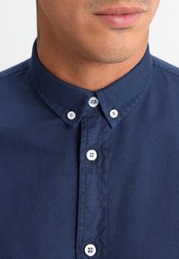 Onderste deel van het gezicht van een man met lichte gezichtsbeharing, gekleed in een donkerblauw overhemd met witte knopen op de kraag en aan de voorkant, dichtgeknoopt.