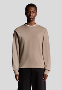 Beige langærmet sweatshirt med rund hals, struktureret stof og subtil logo detalje på brystet. Bæret med mørke bukser.