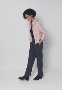 Ljusrosa skjorta knäppt över en svart topp, grå skräddarsydd byxor och svarta stövlar. Håret stylat i mjuka lockar. Enkel bakgrund.