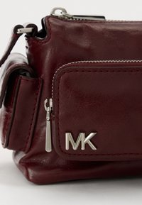 Bordó kožená kabelka se stříbrnými doplňky, zipovou kapsou a embosovaným logem "MK". Obsahuje prošívané detaily a kompaktní design.