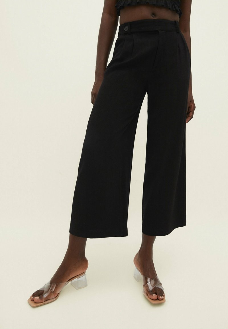 Pantalons noirs à jambes larges en tissu fluide, avec un devant plissé et une fermeture à bouton. Associés à des sandales à talons transparents.