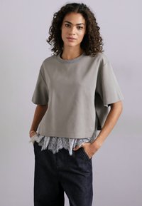 Γκρι κοντό μπλουζάκι με κοντά μανίκια, που διαθέτει δαντελένιο τελείωμα στο στρίφωμα. Η μπλούζα έχει άνετη εφαρμογή και είναι φτιαγμένη από απαλό υλικό.