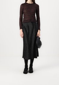 Modell som bär en åtsittande vinröd långärmad topp, kjol i svart satin prydd med glänsande pärlor, svarta strumpbyxor och håller en liten handväska.