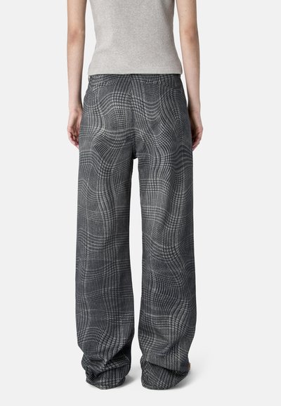 Persona con pantalones sueltos de cuadros grises de cintura alta y una camiseta sin mangas ajustada de color gris claro, de pie frente a un fondo liso.