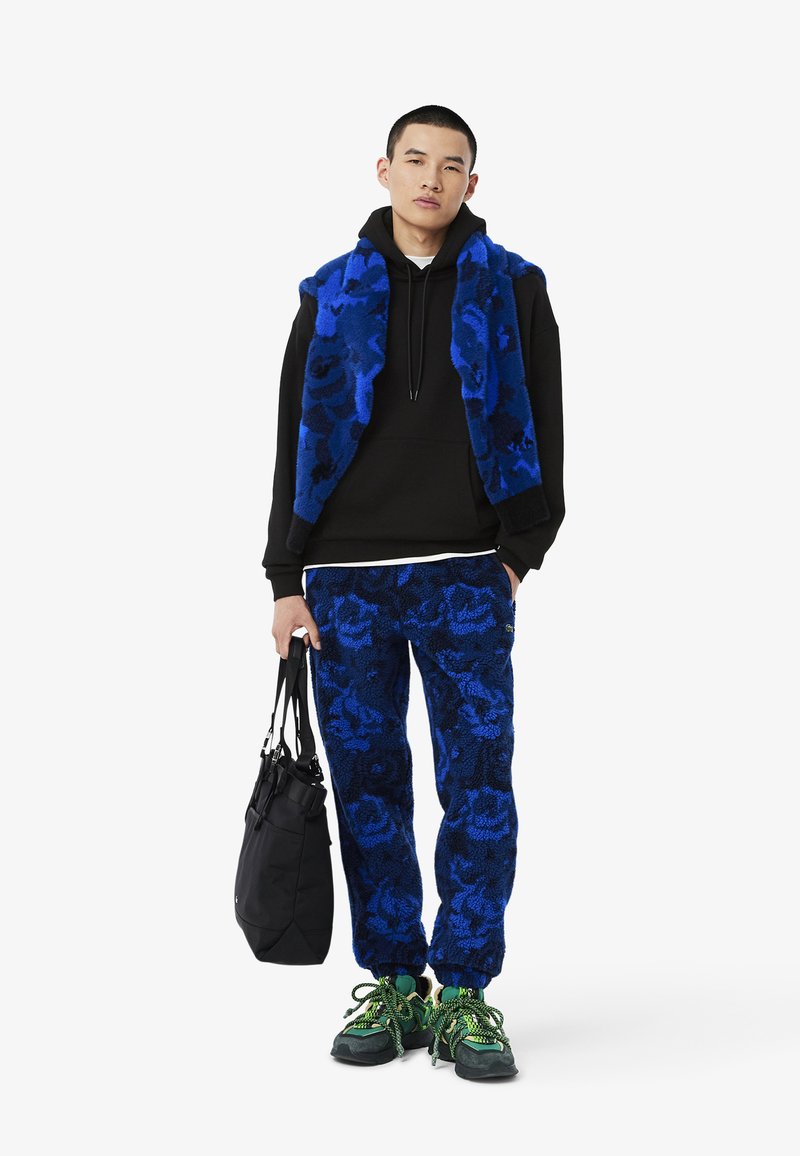 Gilet in pile blu a fantasia sopra una felpa nera, abbinato a pantaloni blu a fantasia e scarpe nere con accenti verdi, mentre porta una borsa nera.