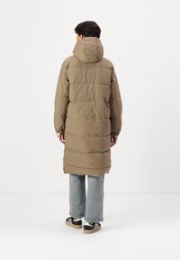 Manteau long rembourré olive avec une capuche, présentant un matelassage horizontal, des poignets élastiques et un ourlet droit. Porté par-dessus un jean clair et des chaussures.