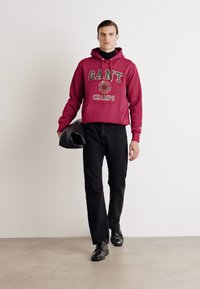 Felpa rossa con cappuccio con la scritta "GANT CHAMPS" in bianco e verde, abbinata a pantaloni neri e scarpe nere, mentre tiene una borsa nera.