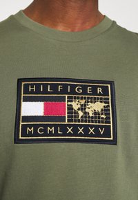 Olivově zelené bavlněné tričko s velkou vyšitou černou nášivkou se zlatým lemem, na které je napsáno "HILFIGER", červený a bílý emblém a mapa světa.