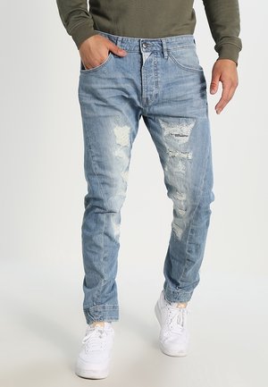 Mann trägt hellblaue, abgenutzte Jeans und weiße Sneaker, mit einer Hand in der Hosentasche und einem olivgrünen Langarmshirt.
