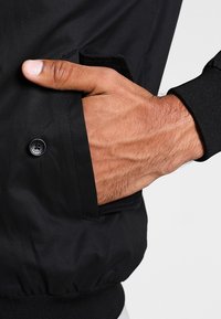 Giacca bomber nera con texture liscia, dotata di una tasca frontale e polsini a coste. Decorata con un bottone nero sulla manica.