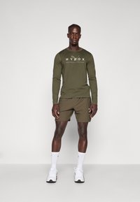 Olijfgroen shirt met lange mouwen en witte tekst, gecombineerd met bijpassende shorts. Model draagt witte sokken en sportschoenen, staande op een effen achtergrond.