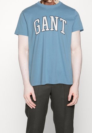 Osoba ubrana w jasnoniebieską koszulkę z logo GANT i ciemne spodnie, stojąca z ramionami swobodnie opuszczonymi wzdłuż ciała na białym tle.