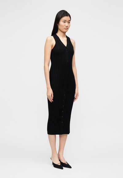 Vestido midi preto com textura canelada, design sem mangas, com frente abotoada e silhueta ajustada. Combinado com sapatilhas pretas pontiagudas.