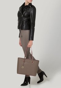 Sac fourre-tout marron avec garniture rose et ferrures argentées. Le sac a des poignées doubles et un pendentif métallique. Le mannequin porte une veste en cuir noire et un pantalon ajusté.