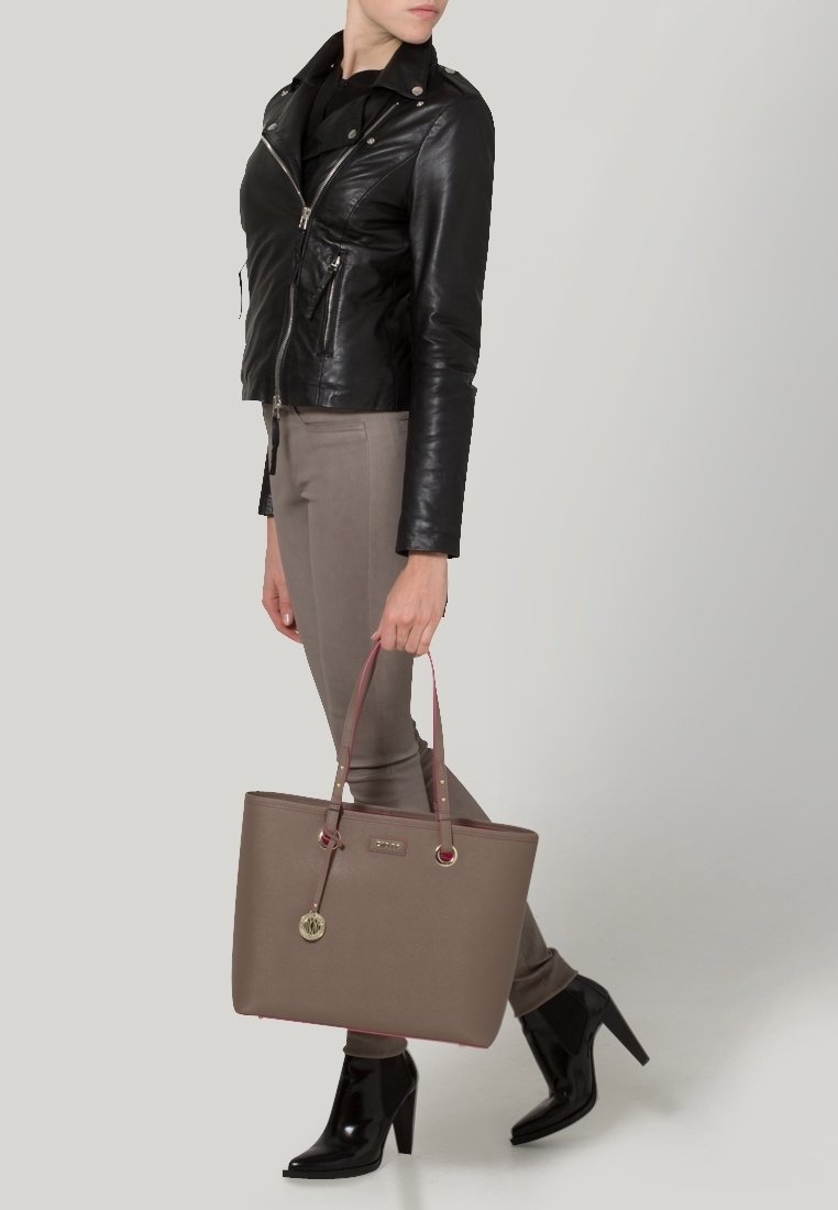 Sac fourre-tout marron avec garniture rose et ferrures argentées. Le sac a des poignées doubles et un pendentif métallique. Le mannequin porte une veste en cuir noire et un pantalon ajusté.