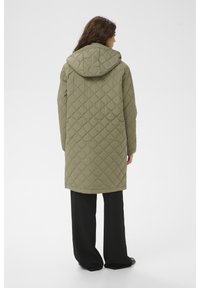 Zaļa pāršūta parka ar kapuci, ar garu griezumu un dimanta rakstu. Valkāta kopā ar melnām platām biksēm, izceļot ērtu dizainu.