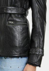 Zwarte leren jas met ritszak, ceintuur in de taille en metalen accenten. Heeft een getextureerde, met bont gevoerde onderrand en mouwdetails.
