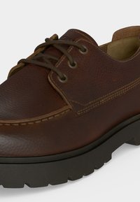 Καφέ δερμάτινο casual παπούτσι με χοντρή μαύρη λαστιχένια σόλα, ραμμένα ραφές και σκούρα καφέ κορδόνια δεμένα μέσα από μεταλλικά πόρπες.