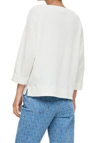 Valkoinen, teksturoidusta materiaalista valmistettu ylisuuri neulepaita, jossa on kolmella neljänneksellä hihat, leveä pääntie ja sivureiät. Tämä yhdistetään vaaleansinisiin, kuviollisiin farkkuihin.