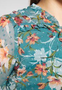 Blouse florale en tissu transparent teal avec des volants, des motifs de fleurs colorées et des points texturés. Accents vifs en orange et rose.