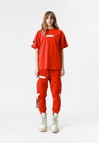 Camiseta de algodón roja con una etiqueta blanca rectangular. Combinada con pantalones rojos que tienen bordados en blanco y un bolsillo lateral con cremallera. Botas blancas con cordones.