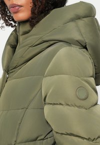 Grønn pufferjakke med quiltet design, med høy krage og logo-patch på ermet. Glatt tekstur og hette.