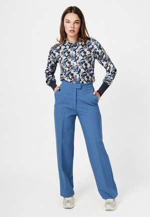 Bloemenprint shirt in marineblauw, crème en bordeaux, met knopen vooraan en geribbelde manchetten; hoge blauwe broek met plooien aan de voorkant.