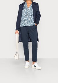 Cappotto trench navy su una blusa floreale blu, abbinato a pantaloni scuri e sneakers bianche. L'outfit include una borsa strutturata.