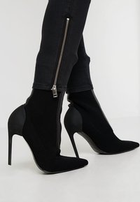 Černé vysoké kozačky na jehlovém podpatku s špičatou špičkou, síťovanou texturou a bočním zipem. Podpatek je zužující, což zlepšuje elegantní siluetu.