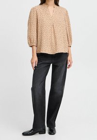 Blouse marron clair à motifs avec des manches trois-quarts et un décolleté en V, associée à un jean droit foncé et des chaussures noires.