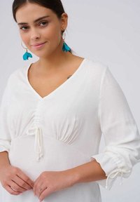 Witte jurk met een V-hals en geplooide details bij de taille. Lange mouwen met elastieken manchetten. Model draagt blauwe bloemenvormige oorbellen.